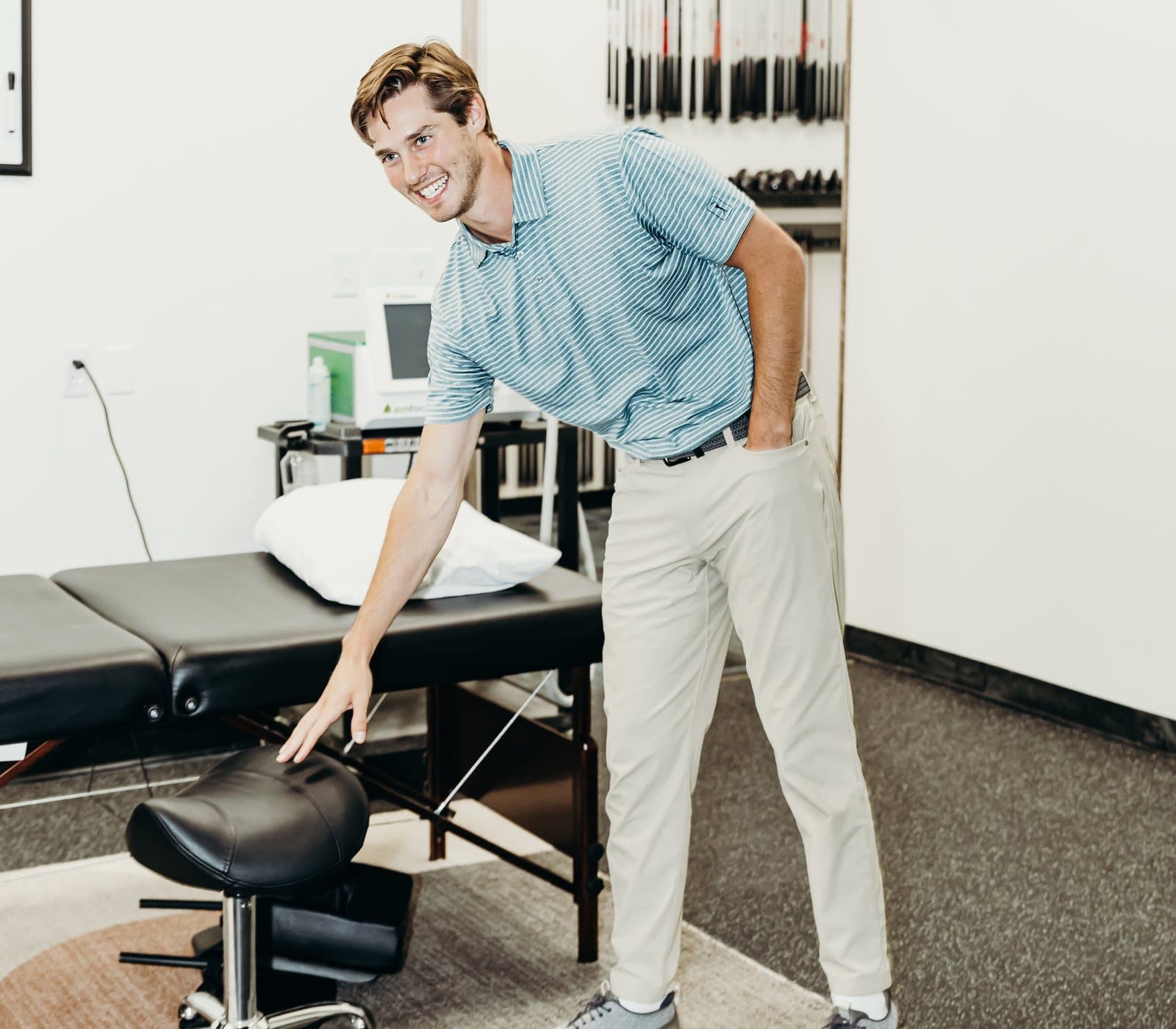 Dr. Taylor Wolf standing beside a treatment table at Elevated Sport and Spine in Salt Lake City, with a rack of golf clubs behind him.